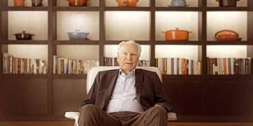 Paul van Zuydam seated in front of cookware and books, reflecting his legacy as Le Creuset’s visionary leader.